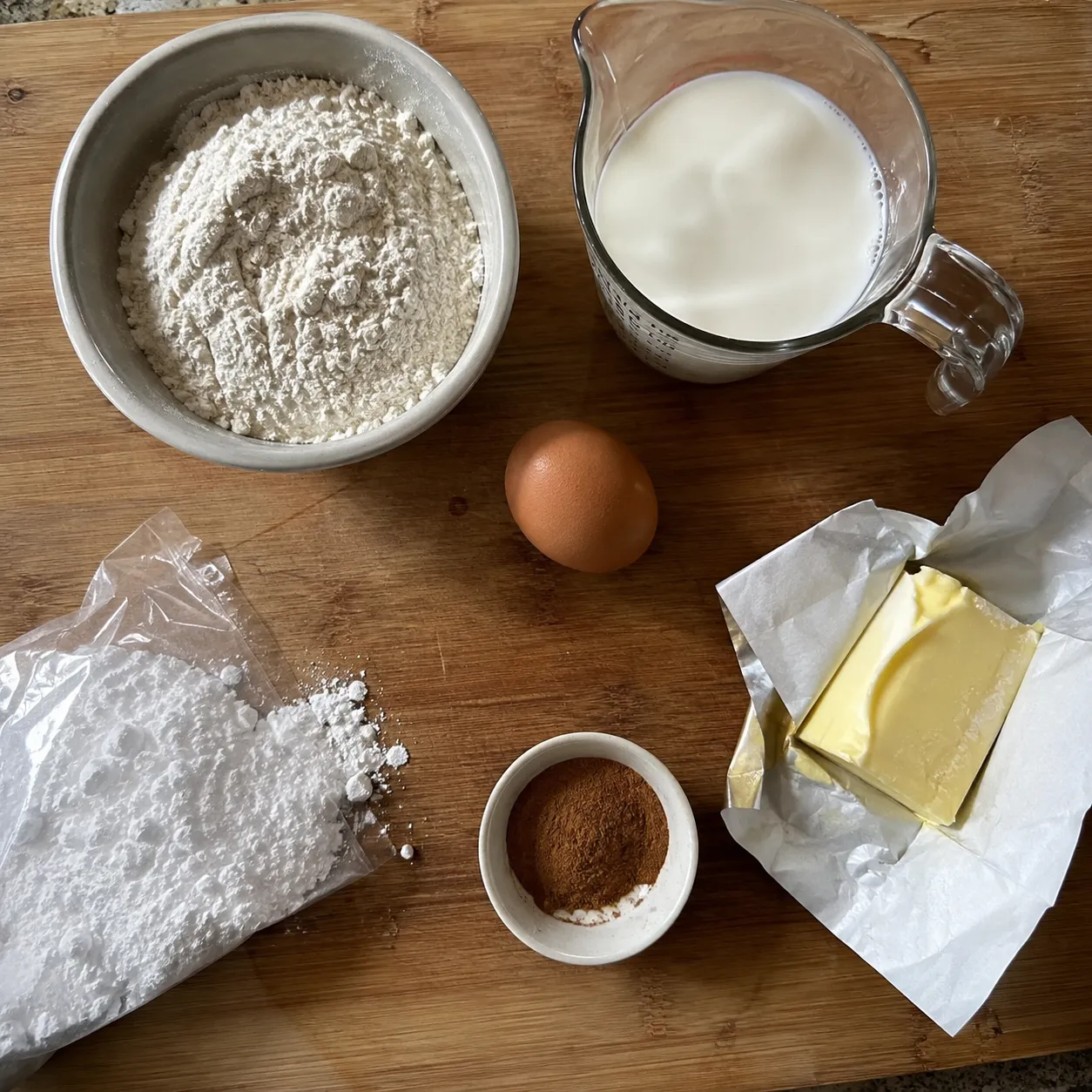 Flour, milk, an egg, butter, ground cinnamon and powdered sugar laid out on a wooden board
