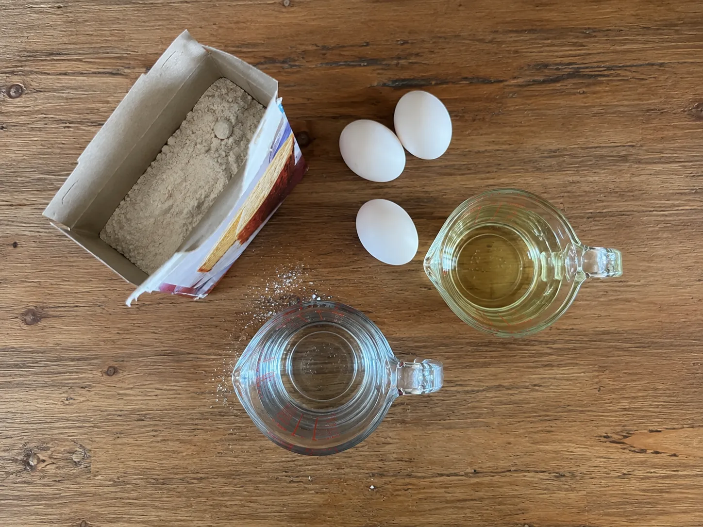 An opened box of cake mix beside three eggs, a jug of oil and a jug of water on a wooden counter