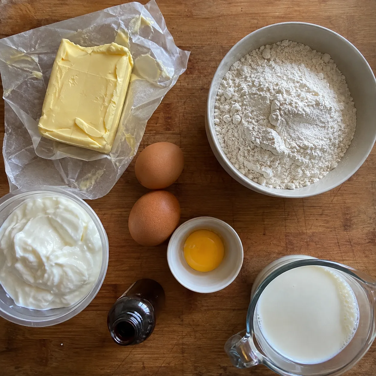 Overhead shot on a wooden board of butter, a bowl of flour, two whole eggs with a third yolk in a small dish, sour cream, vanilla and a jug of milk