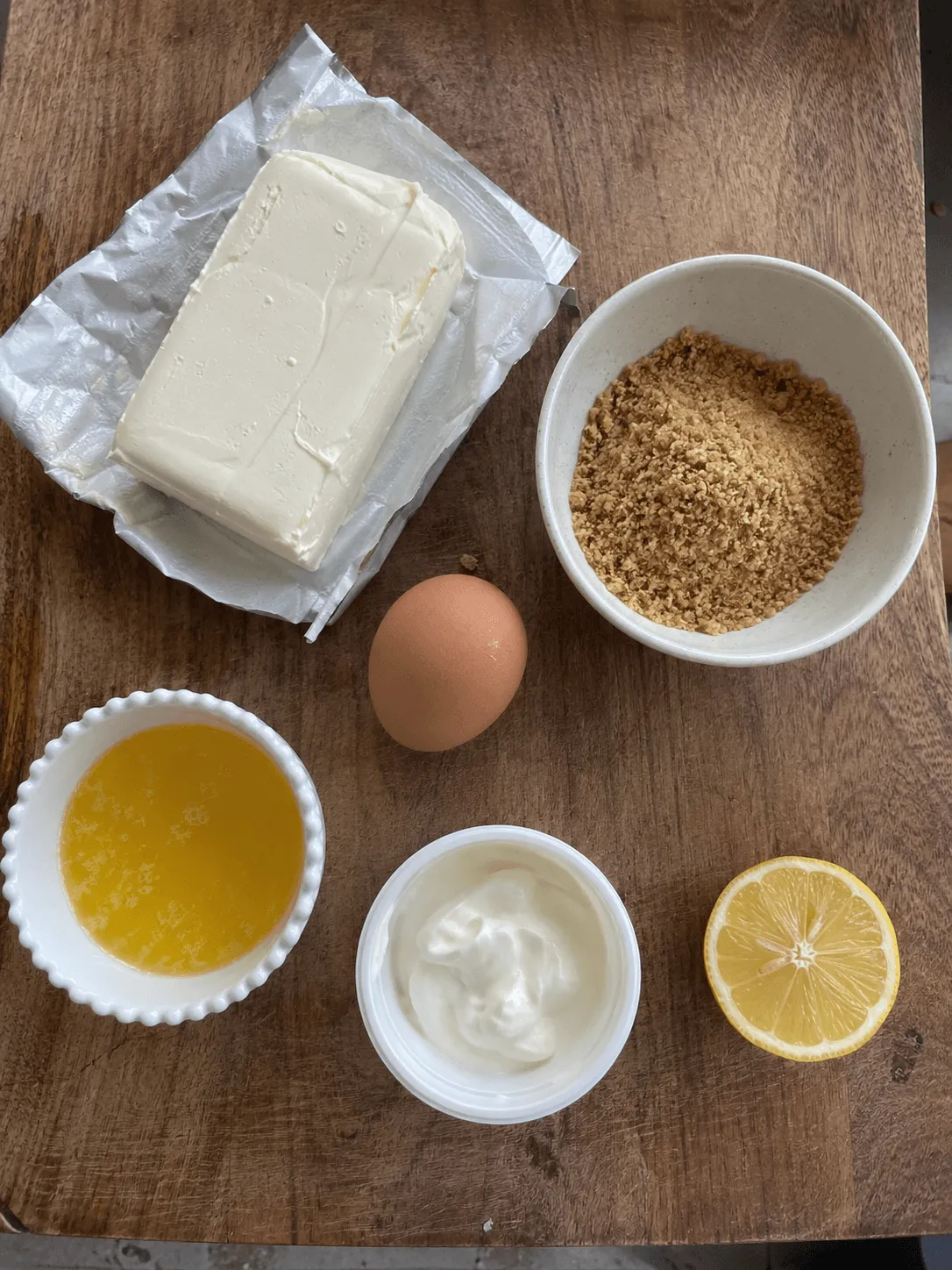 Cream cheese, graham crumbs, melted butter, sour cream, an egg and half a lemon laid out on a wooden board
