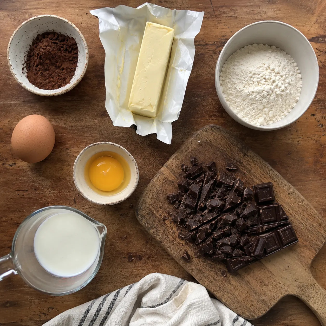 Cocoa, butter, flour, milk, an egg, a separated yolk and chopped dark chocolate on a wooden board