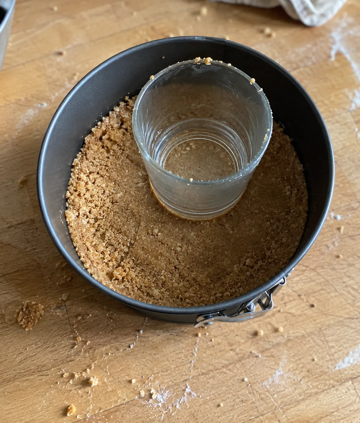 Graham cracker crust being pressed into a springform pan with the flat bottom of a drinking glass