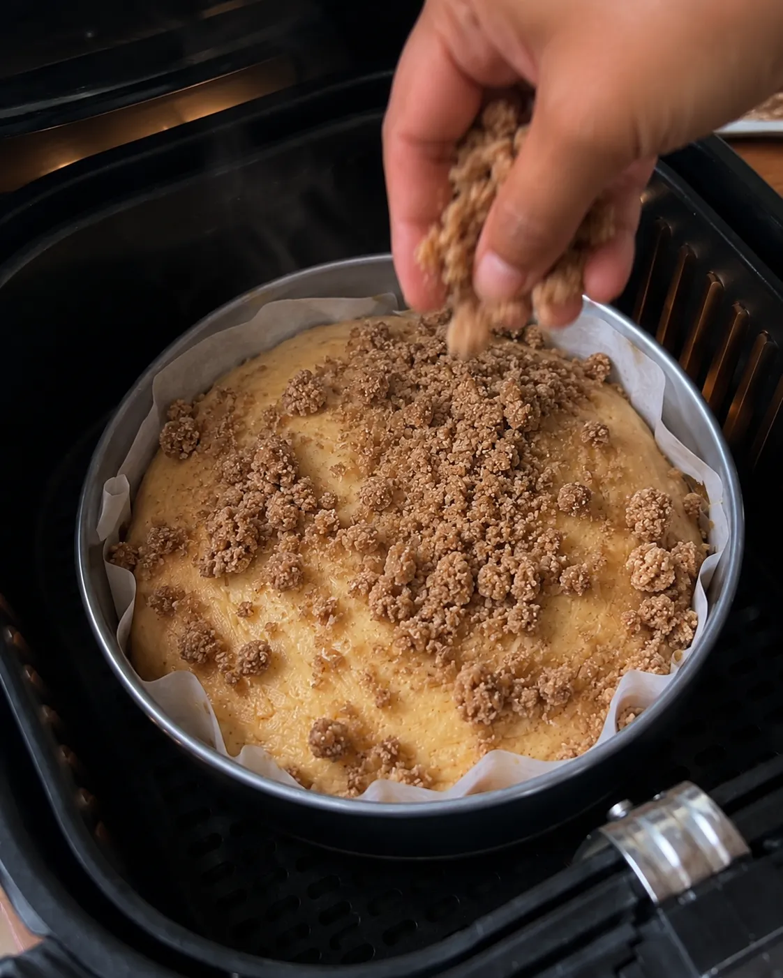A hand scattering cinnamon streusel clumps over the set surface of a part baked cake in an air fryer basket