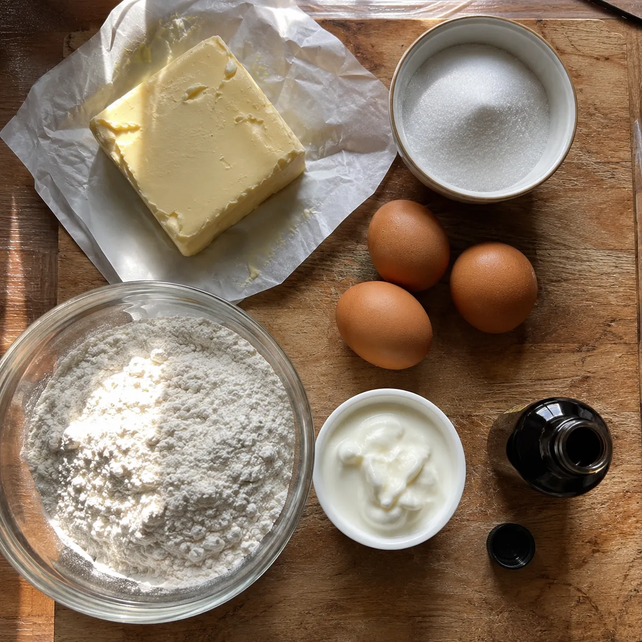 Overhead shot of pound cake ingredients: a block of butter, a bowl of granulated sugar, three eggs, a bowl of flour, a small bowl of sour cream and a bottle of vanilla