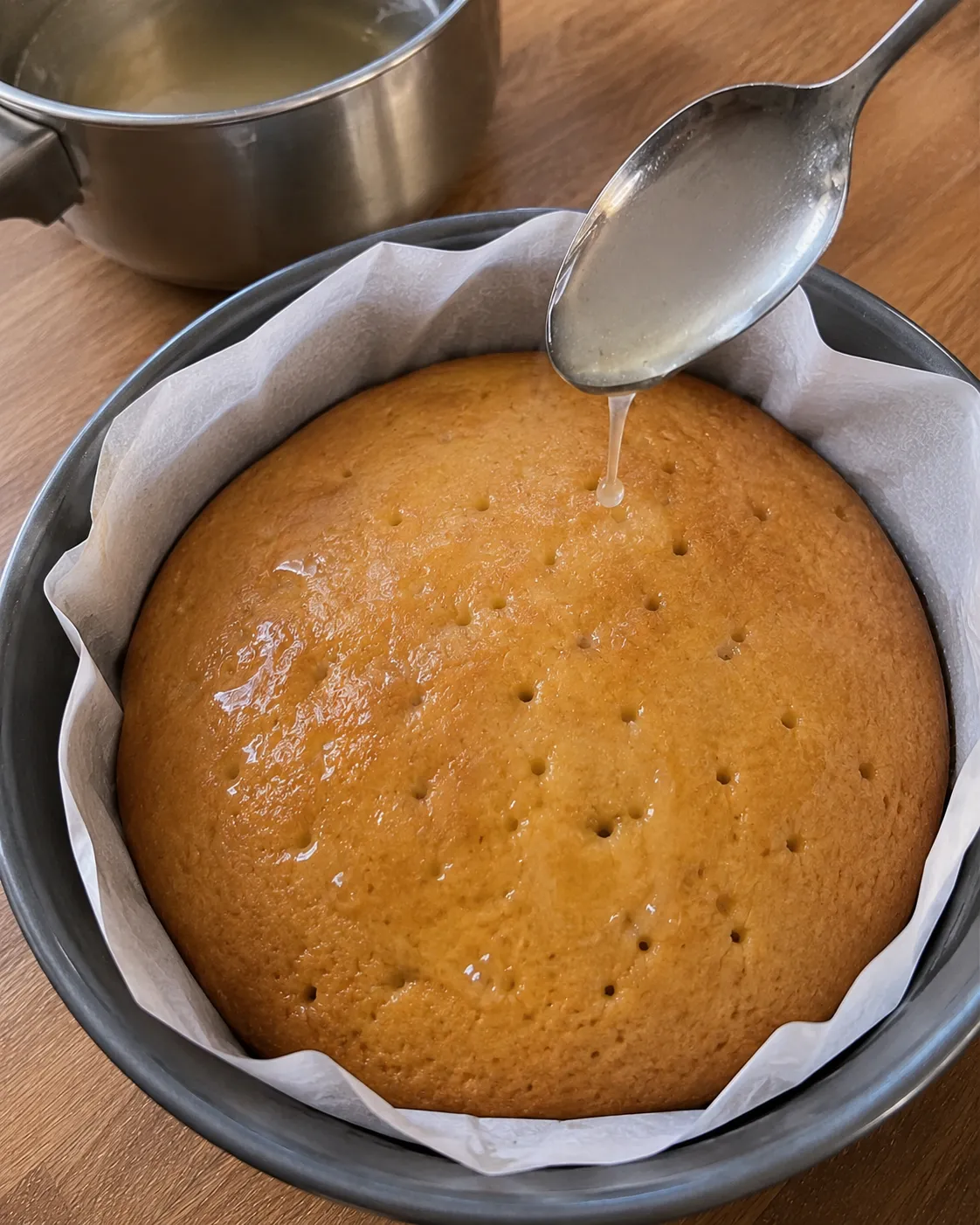 Warm lemon syrup being spooned over a poked cake still in its pan, with skewer holes visible on the surface