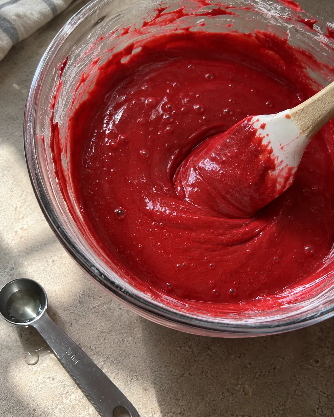 Fully combined red velvet cake batter in a glass bowl, deep even red with no streaks, next to a measuring spoon