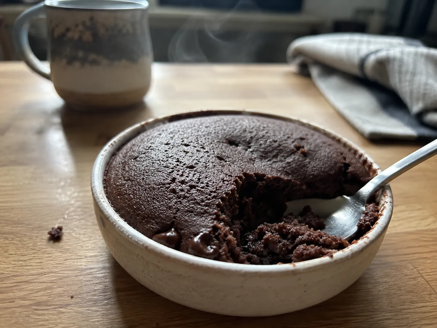 A warm chocolate air fryer mug cake in a white ramekin with a spoon lifting out a piece and a melted chocolate chip on the surface