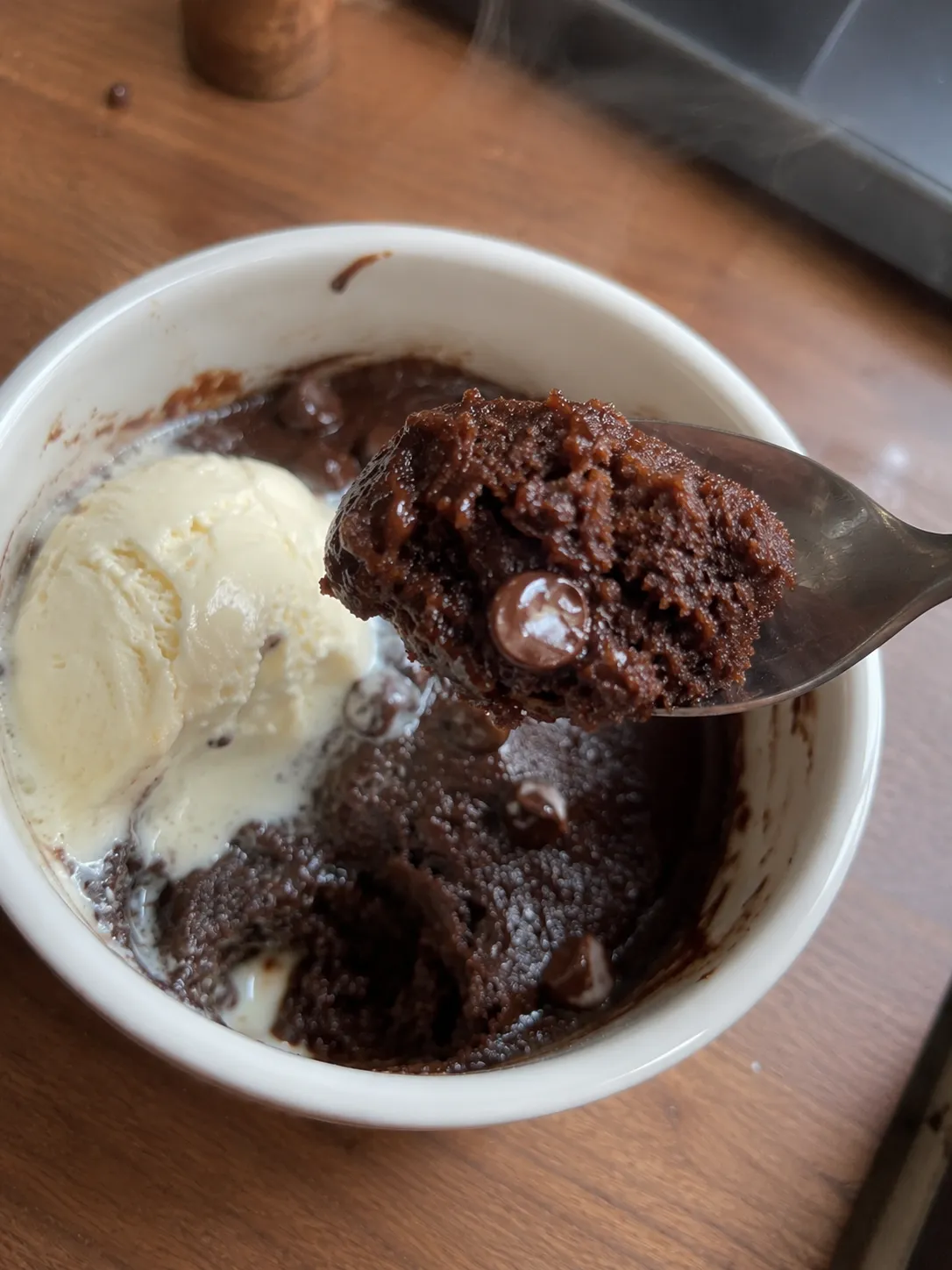 A spoonful of warm chocolate mug cake showing a melted chocolate chip running out of the crumb, with vanilla ice cream in the ramekin