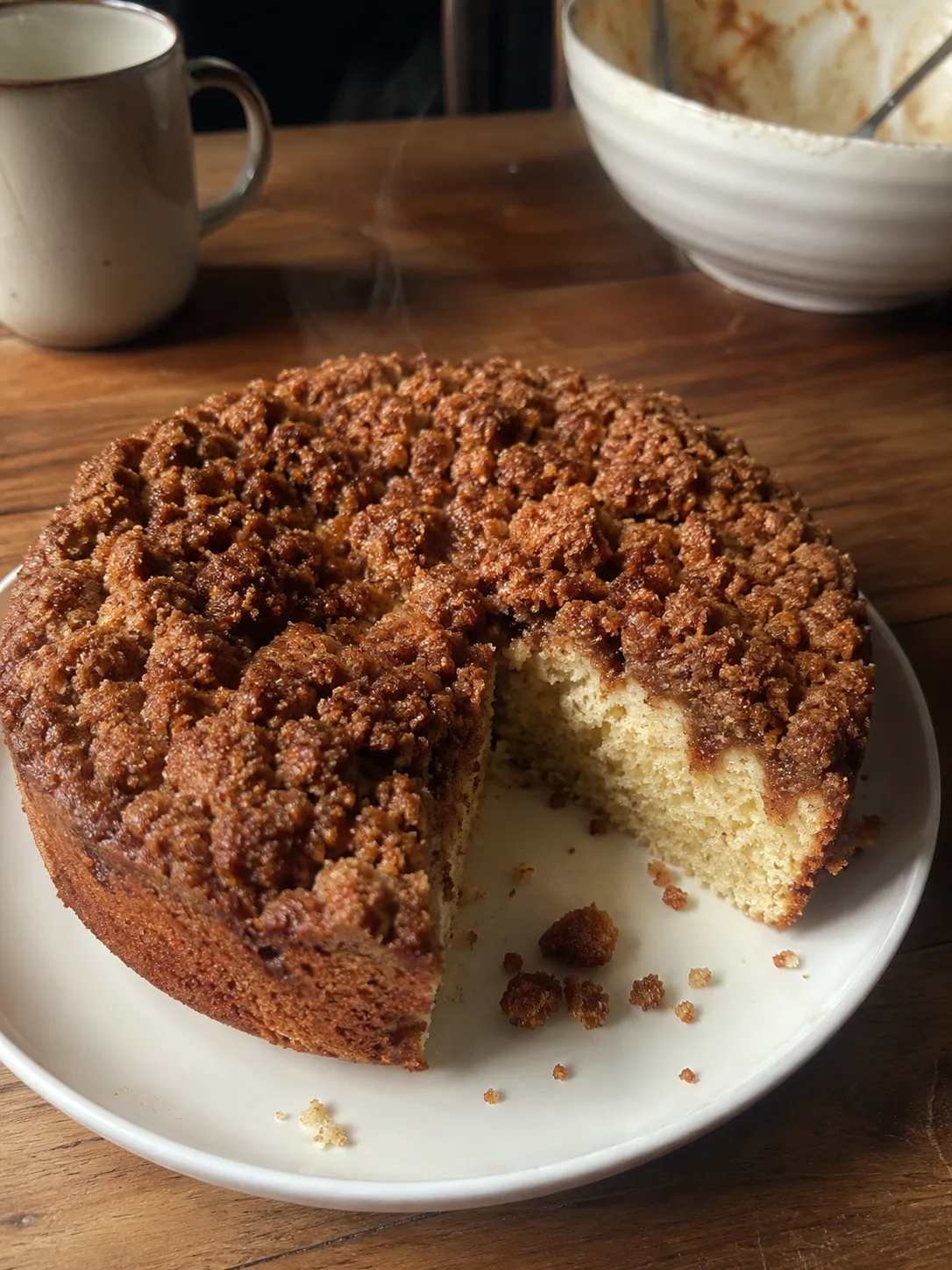 A warm cinnamon streusel coffee cake on a white plate with one wedge cut away, showing a pale tender crumb under a craggy topping