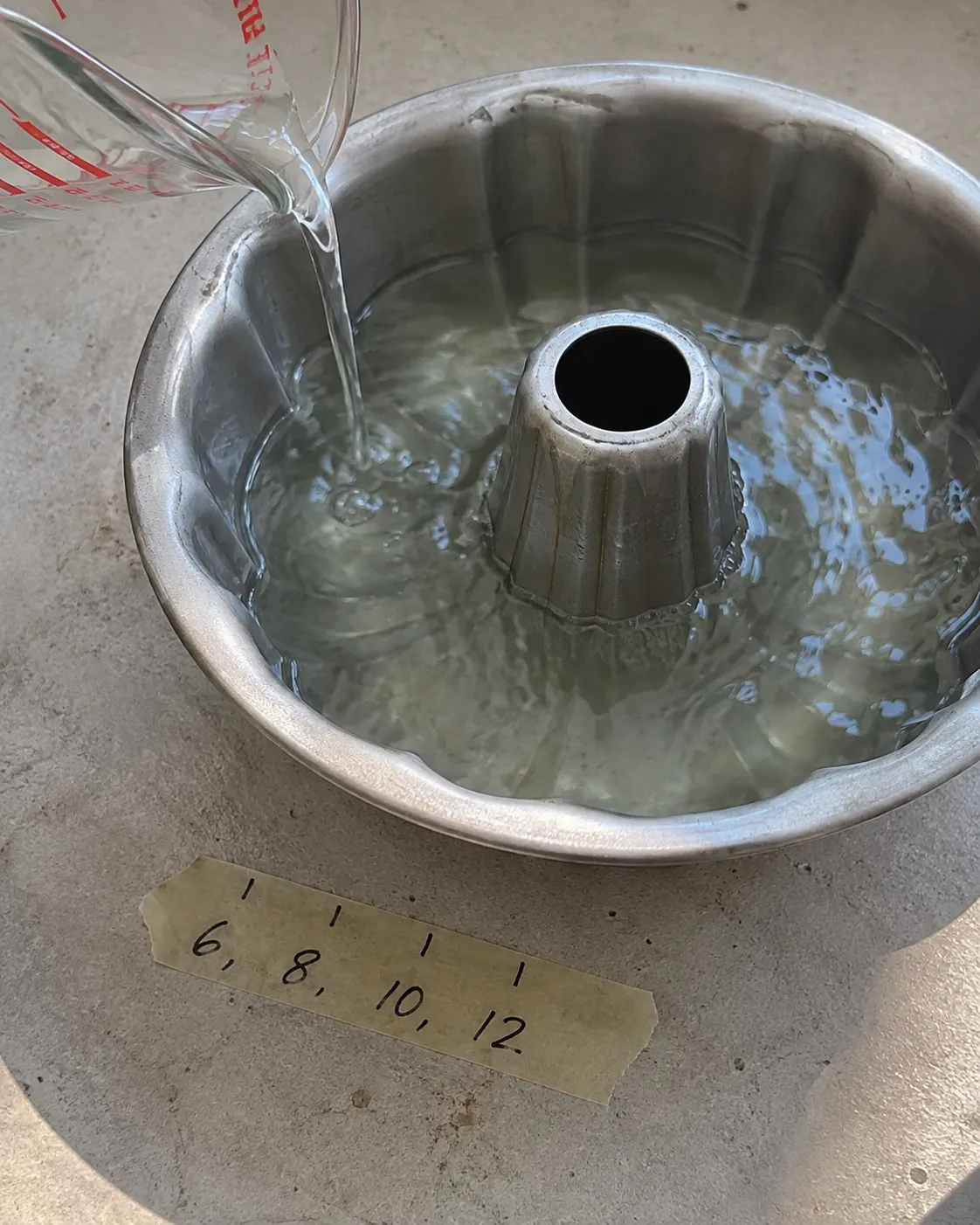 Water being poured from a glass measuring cup into a fluted bundt pan, with a strip of masking tape beside it marked 6, 8, 10, 12