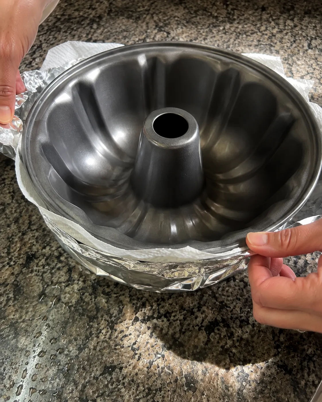 Two hands wrapping a band of foil with a wet paper towel inside it around the outside of an empty fluted tube pan