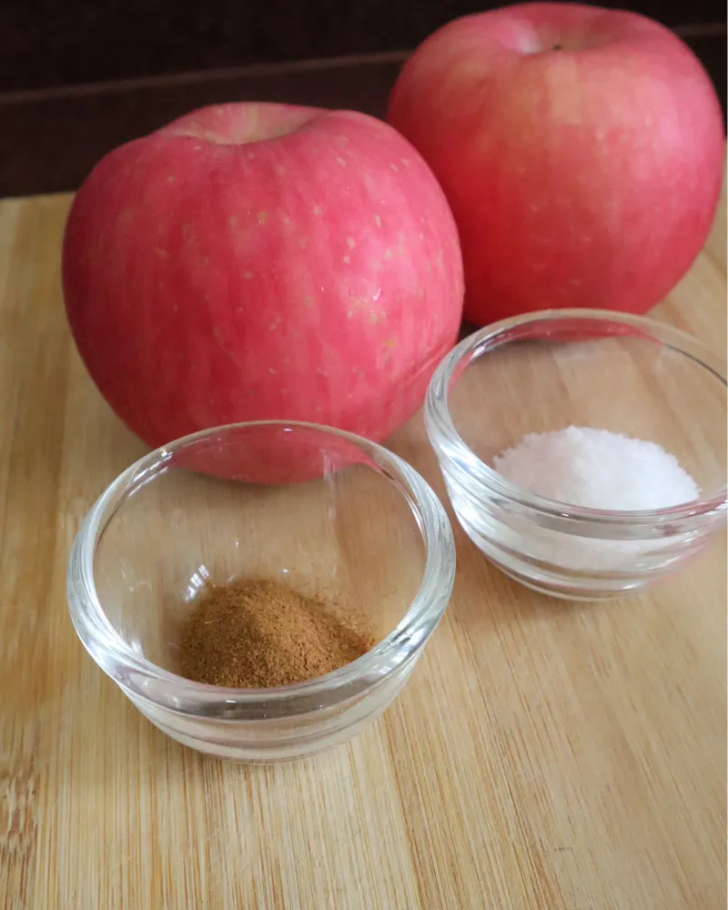 Two red apples on a bamboo board beside small glass bowls of ground cinnamon and sugar