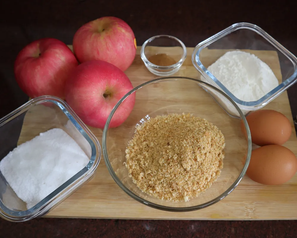 Apples, ground cinnamon, flour, sugar, breadcrumbs and two eggs laid out on a wooden board