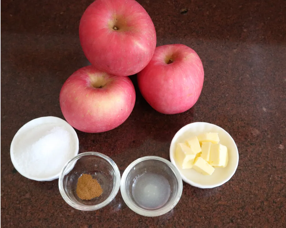 Whole apples with small bowls of sugar, ground cinnamon, water and cubed butter on a counter