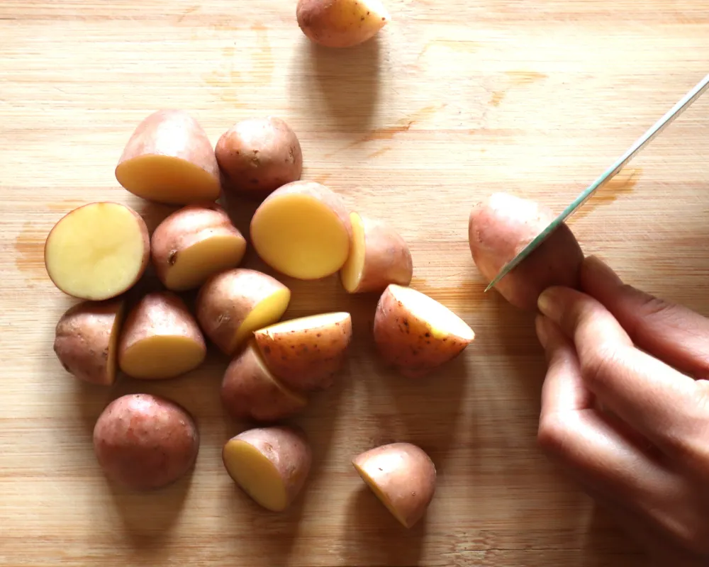 A hand cutting baby potatoes on a wooden board, some halved and some quartered, with the pale waxy flesh showing