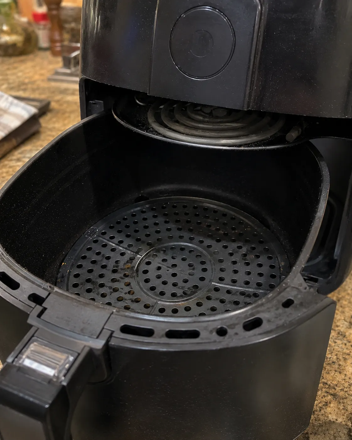 An open basket air fryer showing the perforated crisper plate below and the coiled heating element above