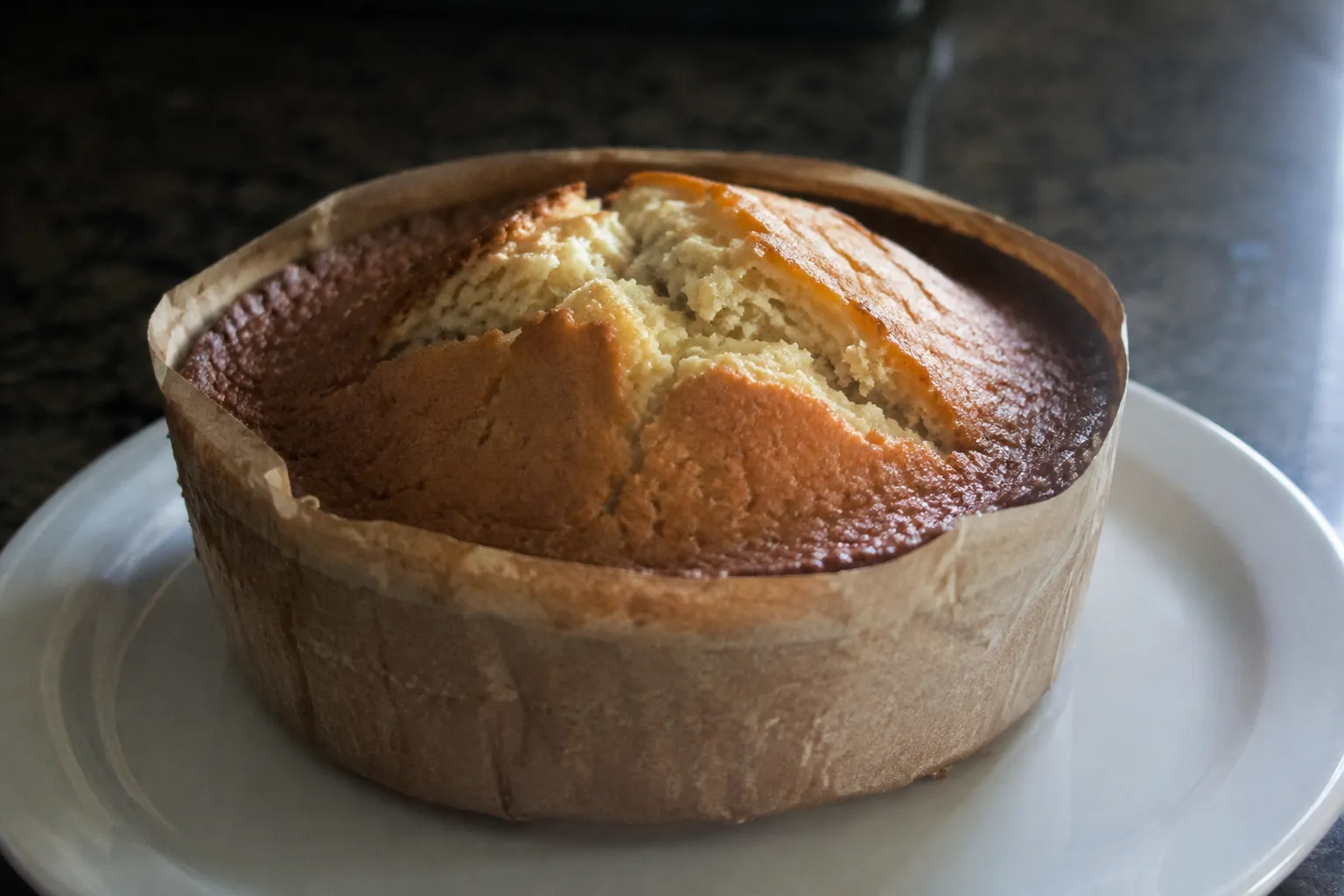 A cake in a parchment collar risen higher in the middle with a split across the top and a dark rim