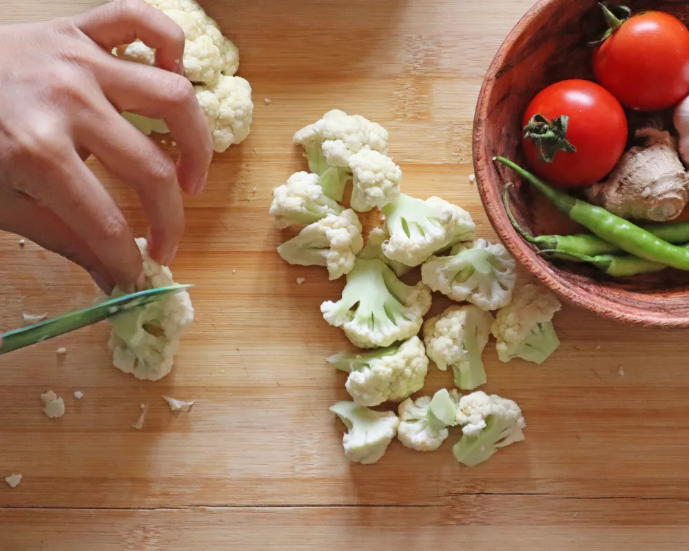 A hand cutting a cauliflower head into florets on a wooden board beside a bowl of tomatoes, green chilies and ginger