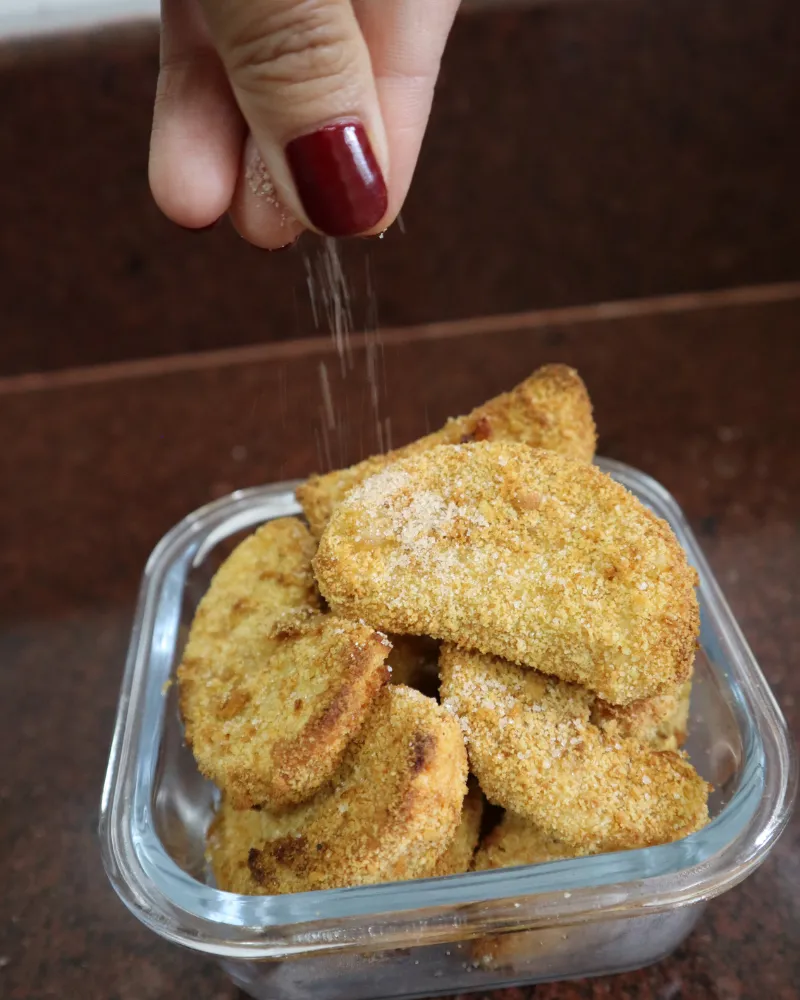 A hand sprinkling cinnamon sugar over hot breaded apple fries in a glass dish