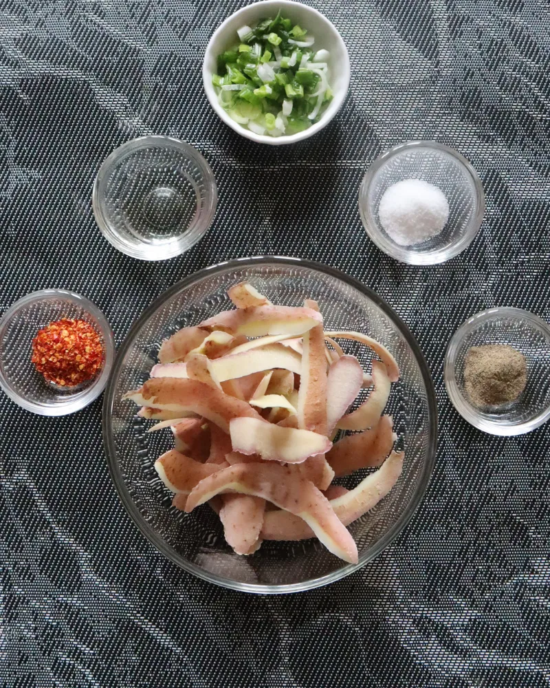 A glass bowl of raw potato peels surrounded by small bowls of scallions, oil, salt, chili flakes and black pepper