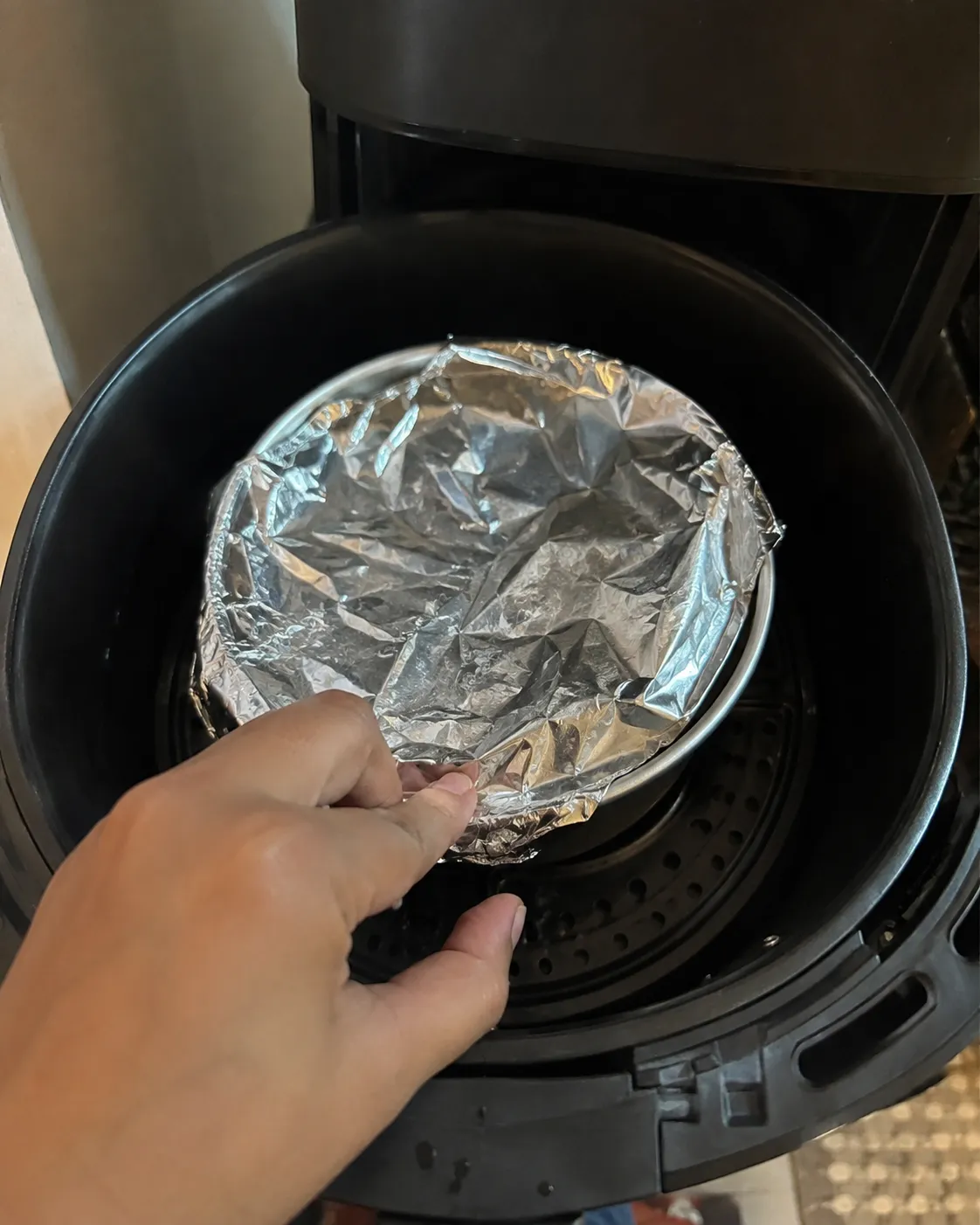 A hand tucking a sheet of foil loosely over a round cake tin inside a black air fryer basket