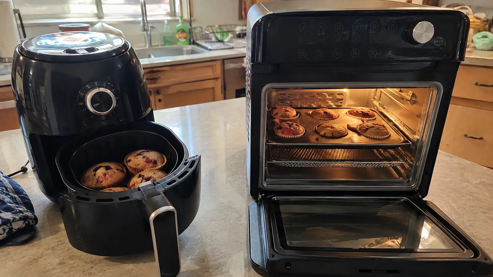 A basket air fryer with muffins beside an oven-style air fryer holding a tray of cookies