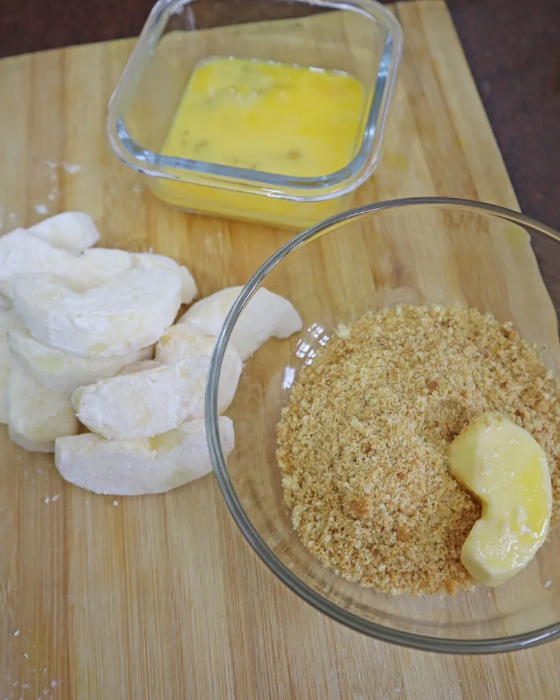An egg-dipped apple piece being pressed into a bowl of breadcrumbs beside flour-coated pieces and a dish of beaten egg