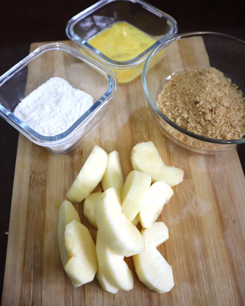 Cut apple pieces on a board in front of three bowls holding flour, beaten egg and cinnamon breadcrumbs