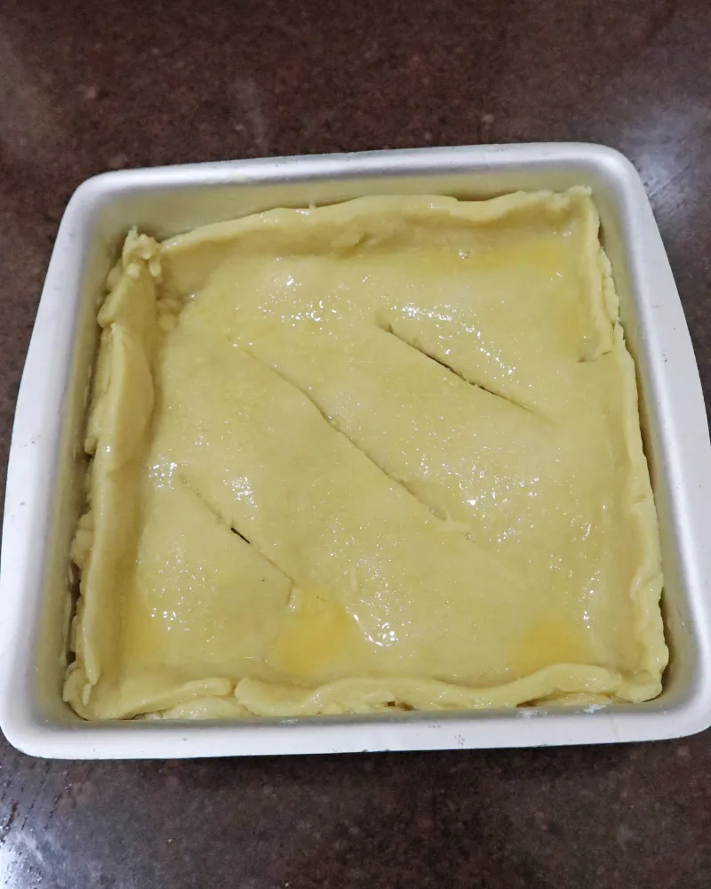 An assembled double crust apple pie in a square metal pan, egg washed with three vents cut in the lid