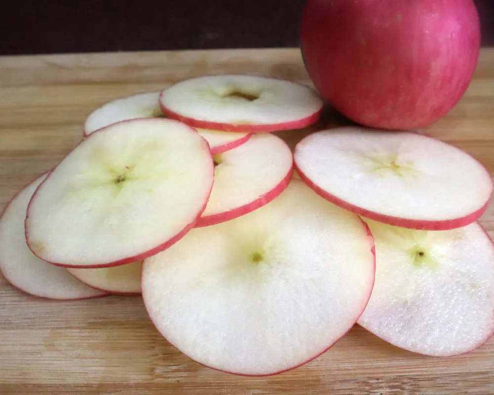 Thin raw apple slices piled on a wooden board beside a whole red apple