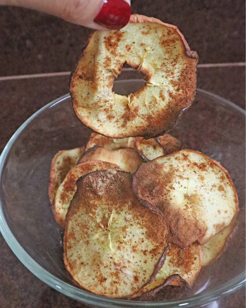 A hand holding one crisp apple chip above a glass bowl of air fryer apple chips