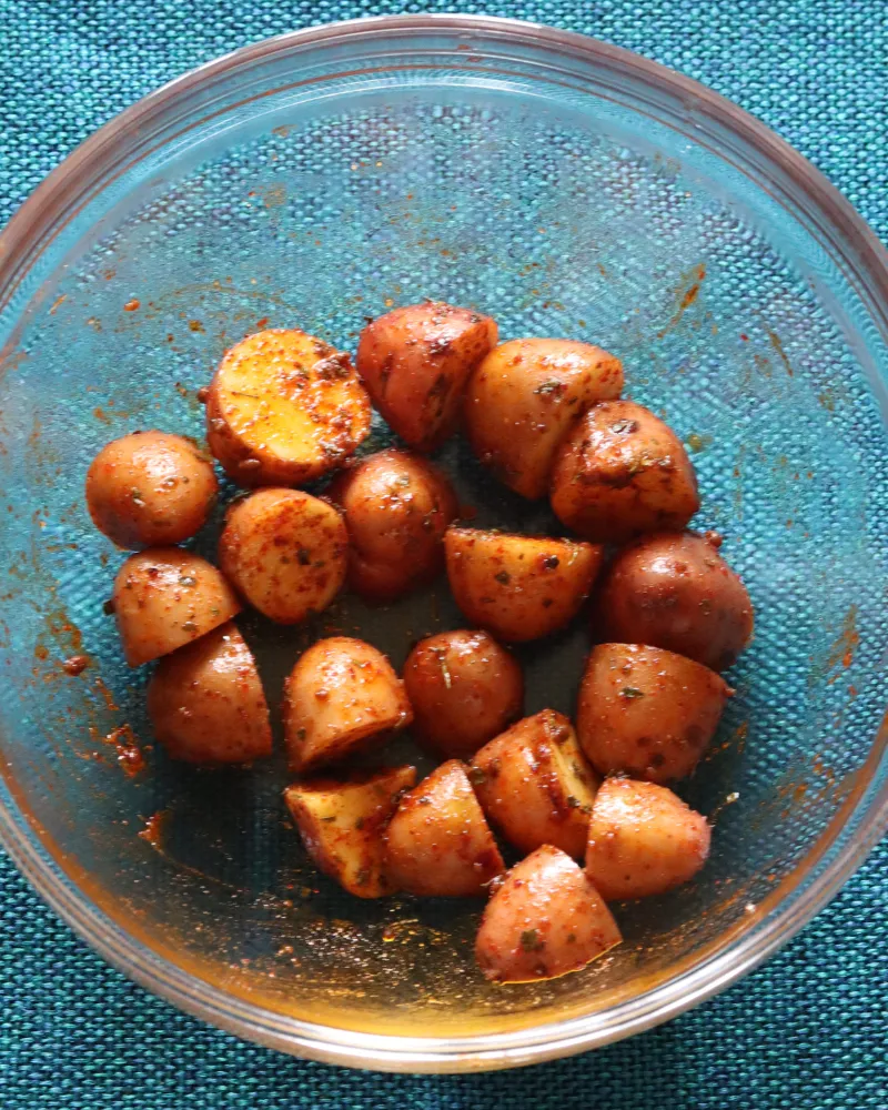Cut baby potatoes tossed with oil and red seasoning in a glass bowl, every surface coated and glossy