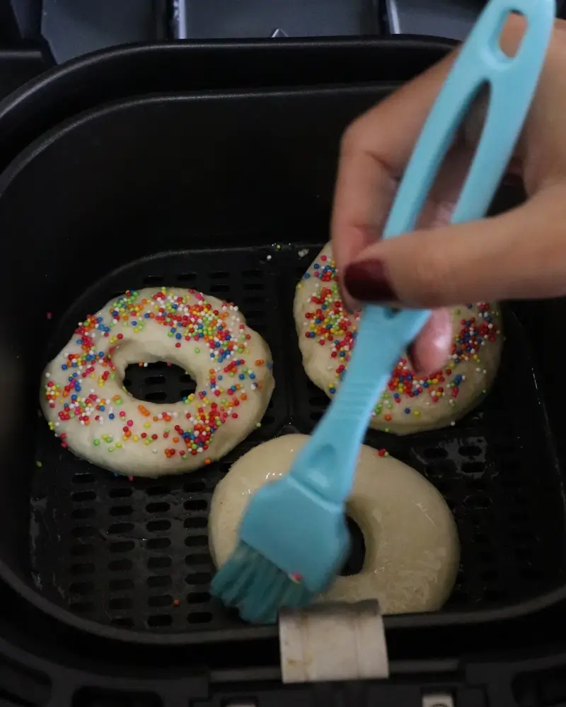 A silicone pastry brush coating a raw vegan donut with melted coconut oil in the air fryer basket