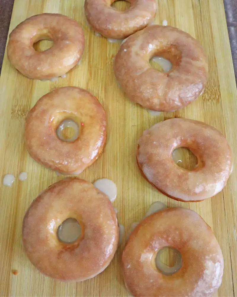 Seven glazed vegan donuts set out on a wooden board with glaze pooling underneath
