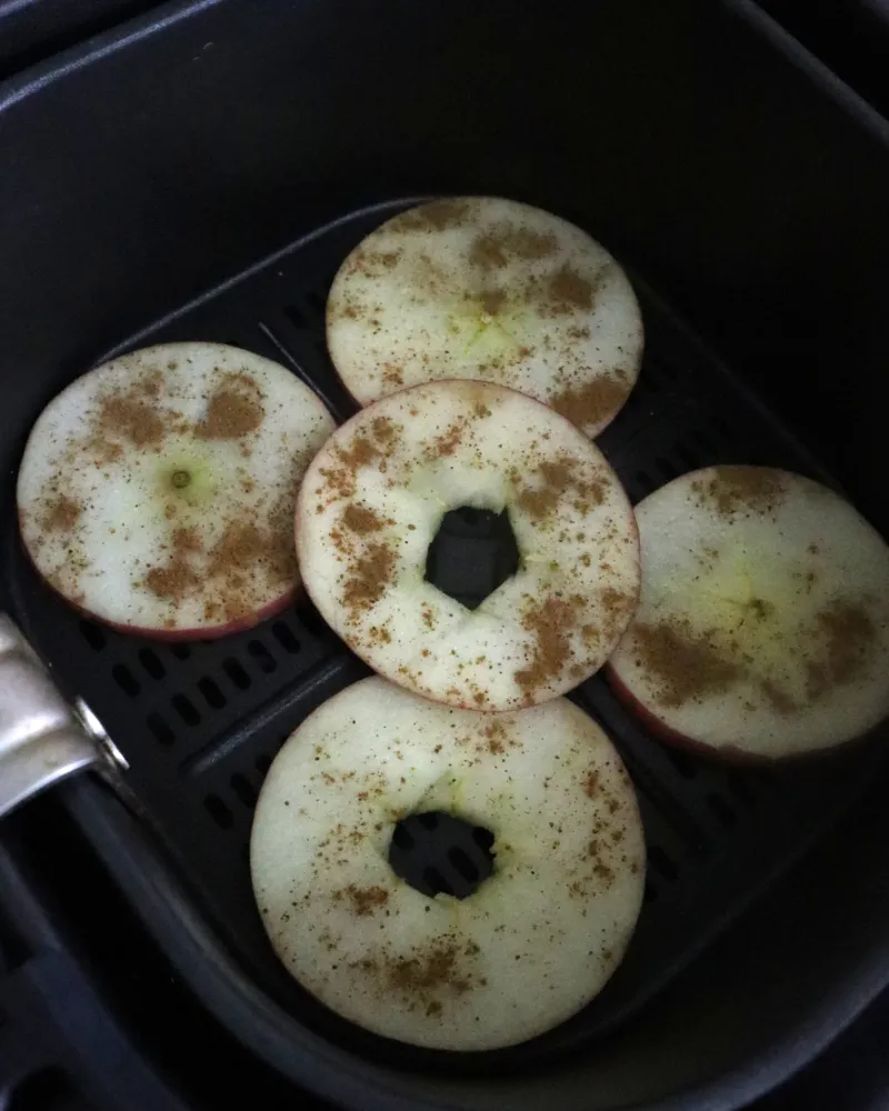 Five raw cinnamon dusted apple rings arranged in a single layer in an air fryer basket