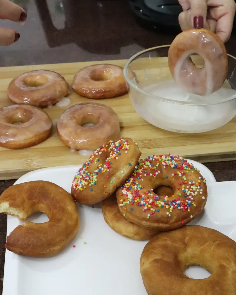 A hand dipping a vegan donut into a glass bowl of white glaze, with glazed and sprinkled donuts alongside