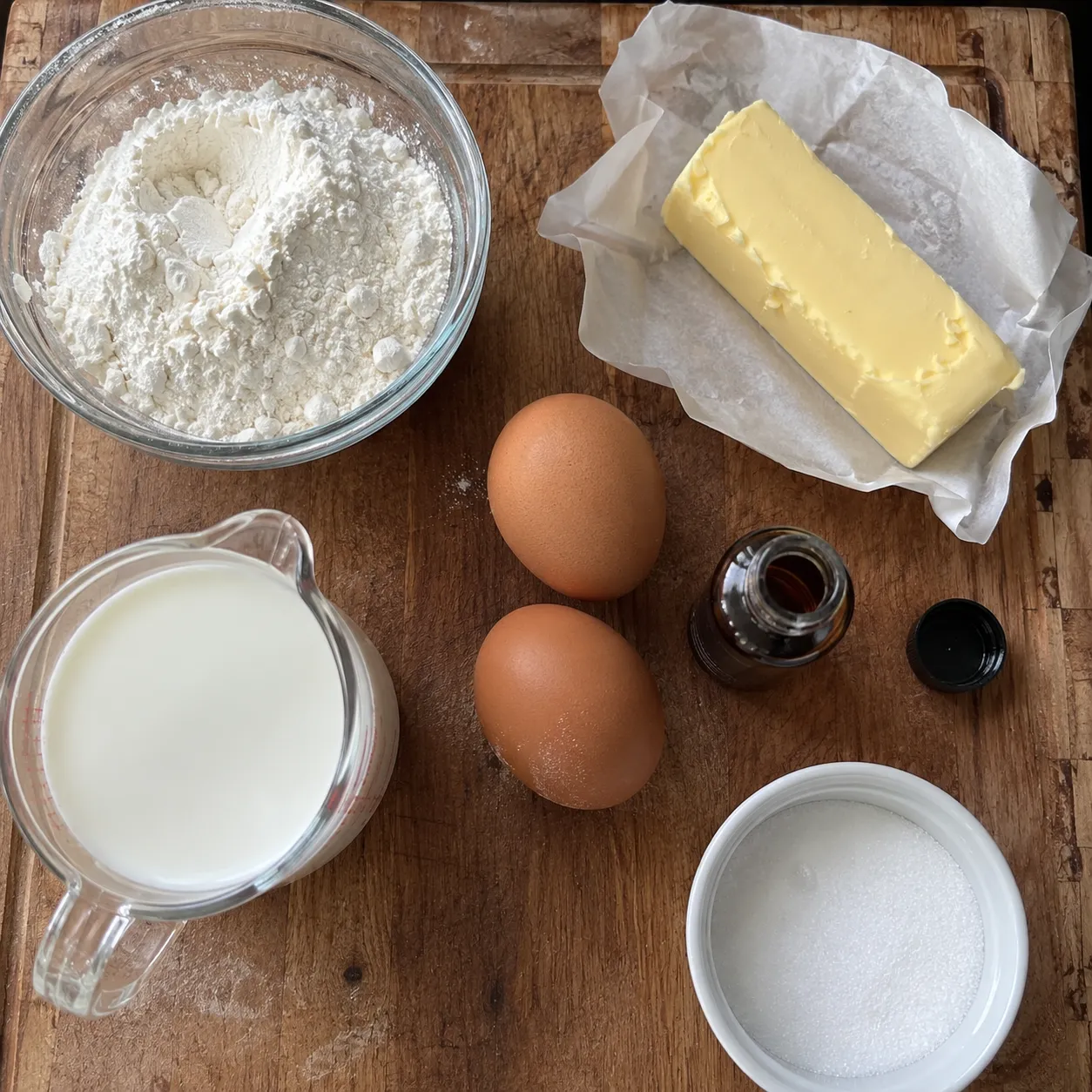 Flour, butter, two eggs, milk, vanilla extract and sugar laid out on a wooden board