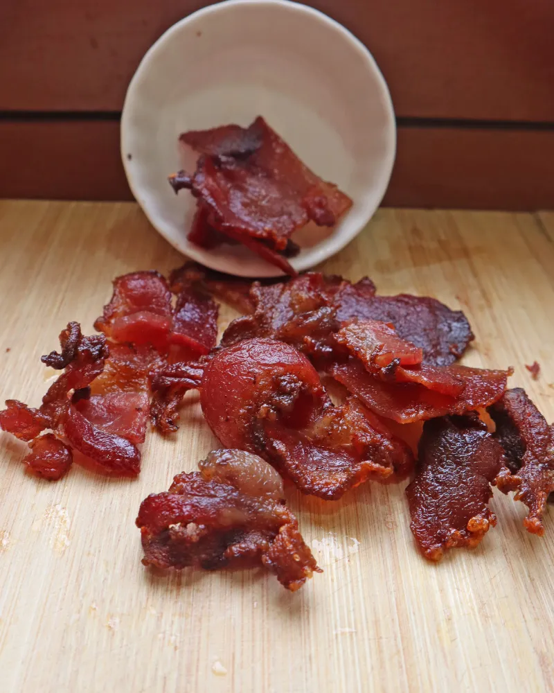 Cooked air fryer bacon spilling from a tipped white bowl onto a wooden board