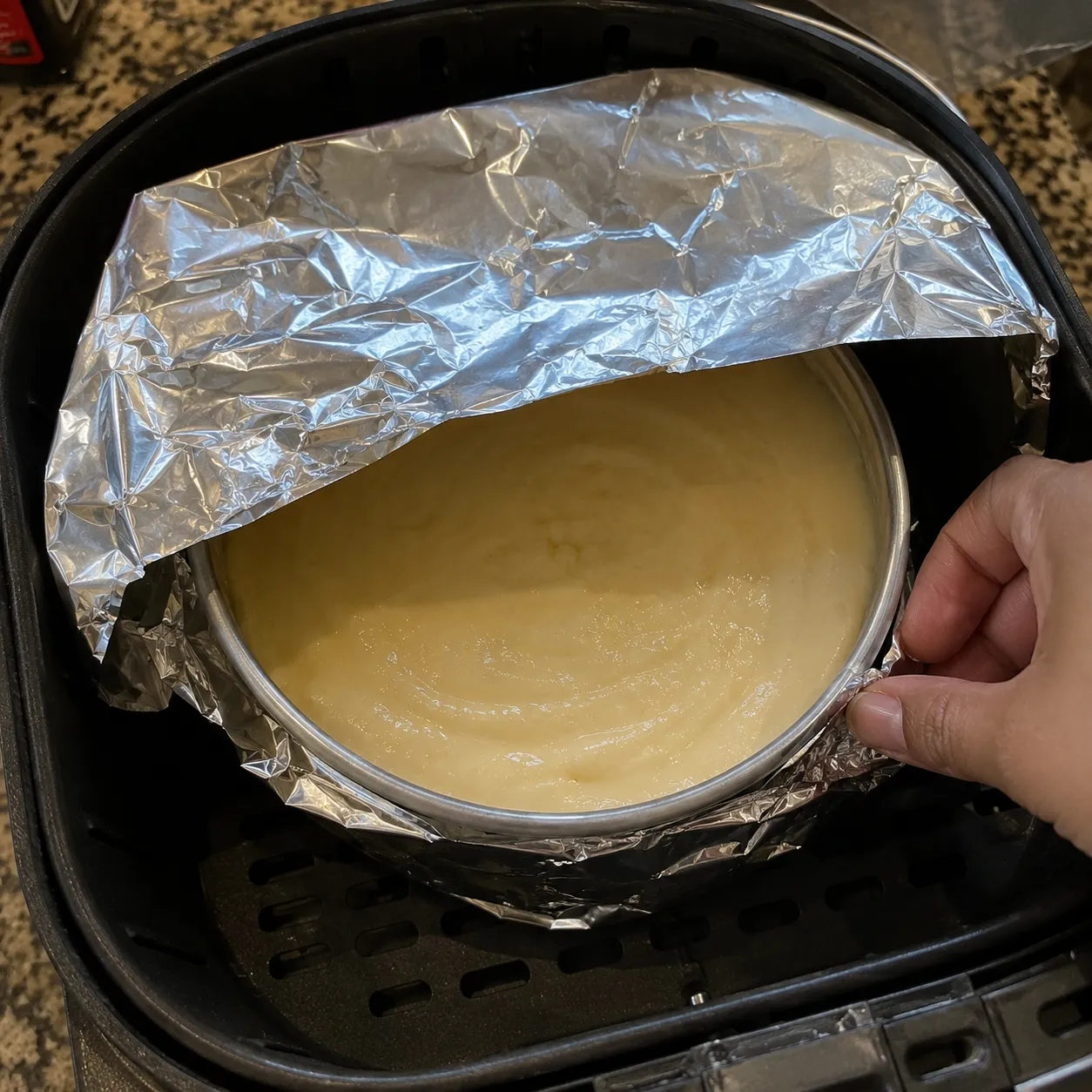 A hand lowering a foil-covered tin of vanilla cake batter into an air fryer basket
