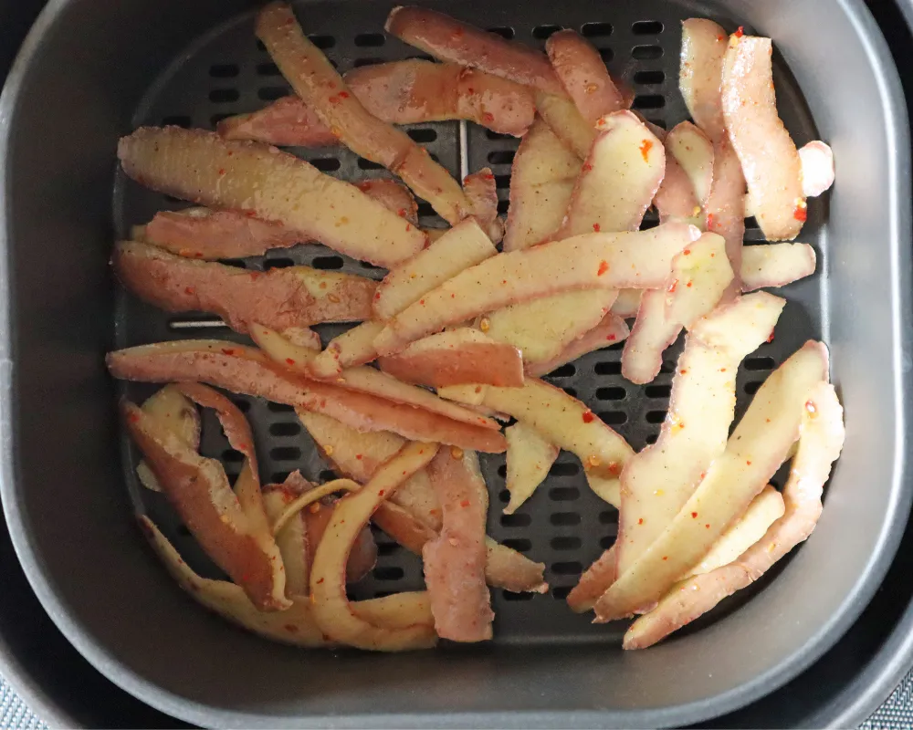 Raw oiled potato peels spread in a single layer across an air fryer basket