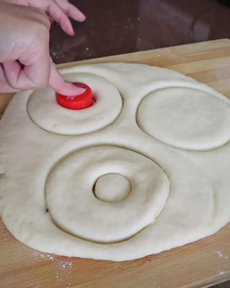 A hand pressing a red bottle cap through rolled vegan donut dough to cut out the center hole