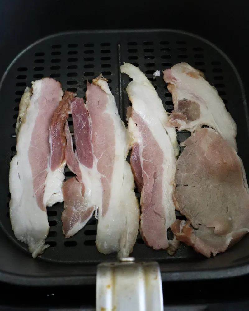 Raw thick cut bacon strips laid side by side across an air fryer basket before cooking