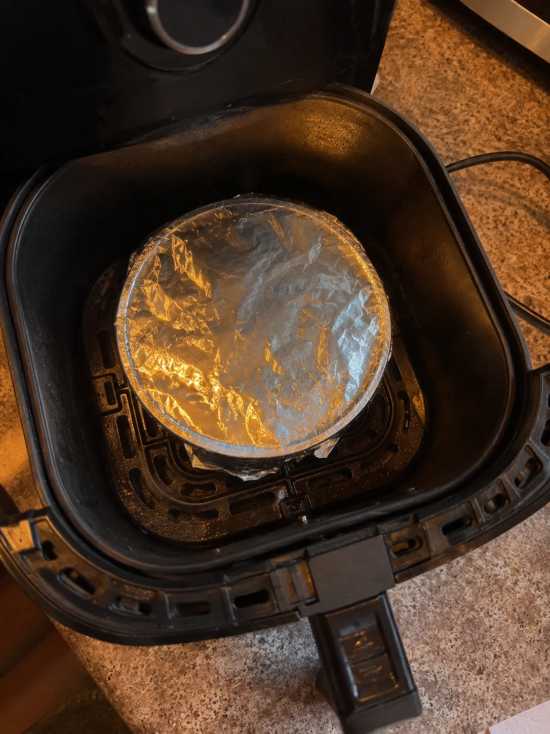 A cake tin covered with foil sitting in an air fryer basket, the foil tucked down around the rim