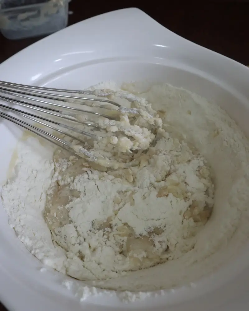 A whisk working plant milk and melted coconut oil into flour in a white mixing bowl