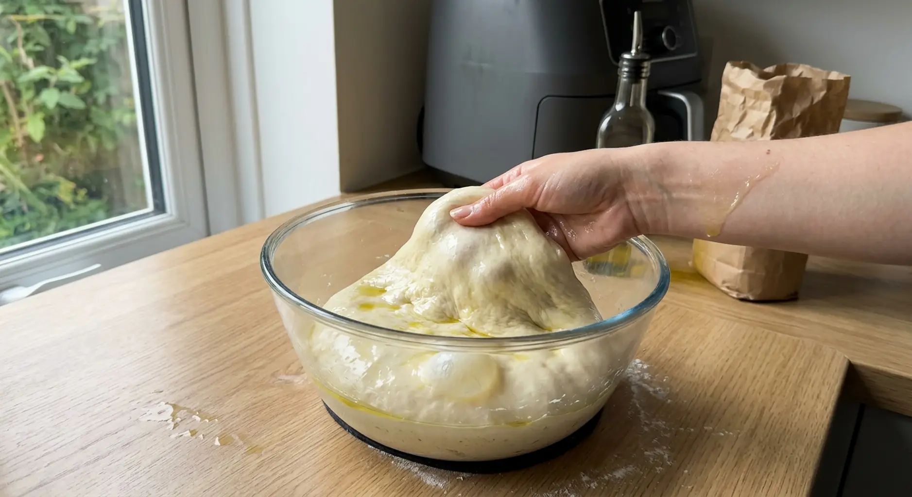 An oiled hand lifting and folding wet focaccia dough over itself inside a glass bowl
