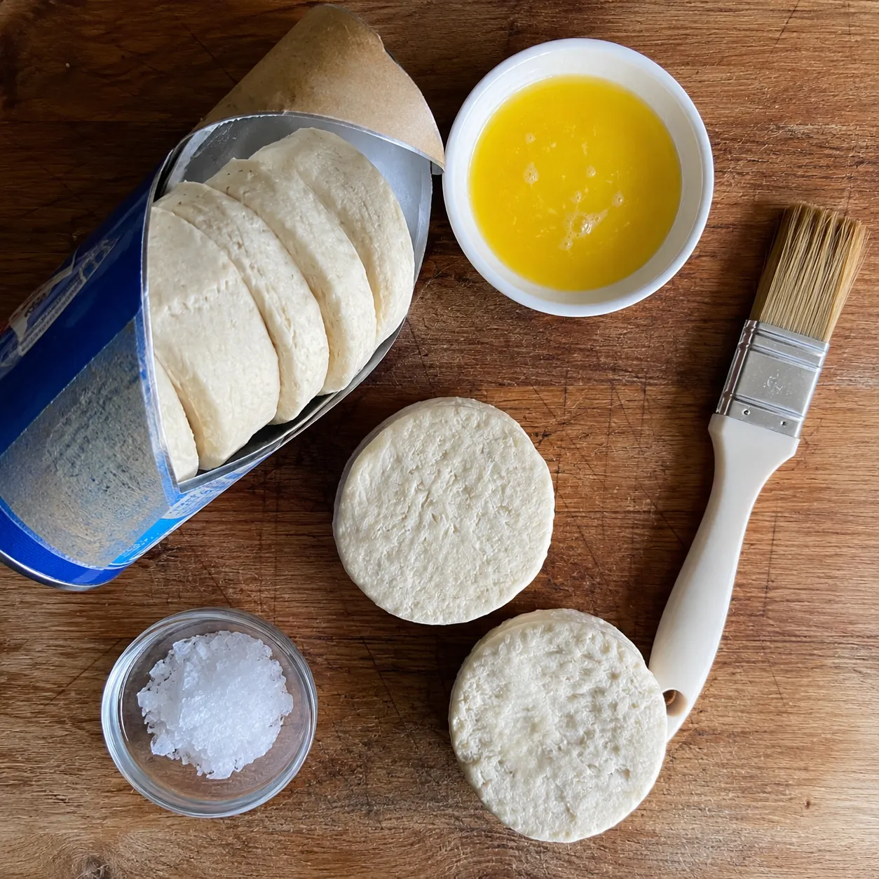 A can of biscuits opened on a wooden board with separated rounds, melted butter, a pastry brush and flaky salt