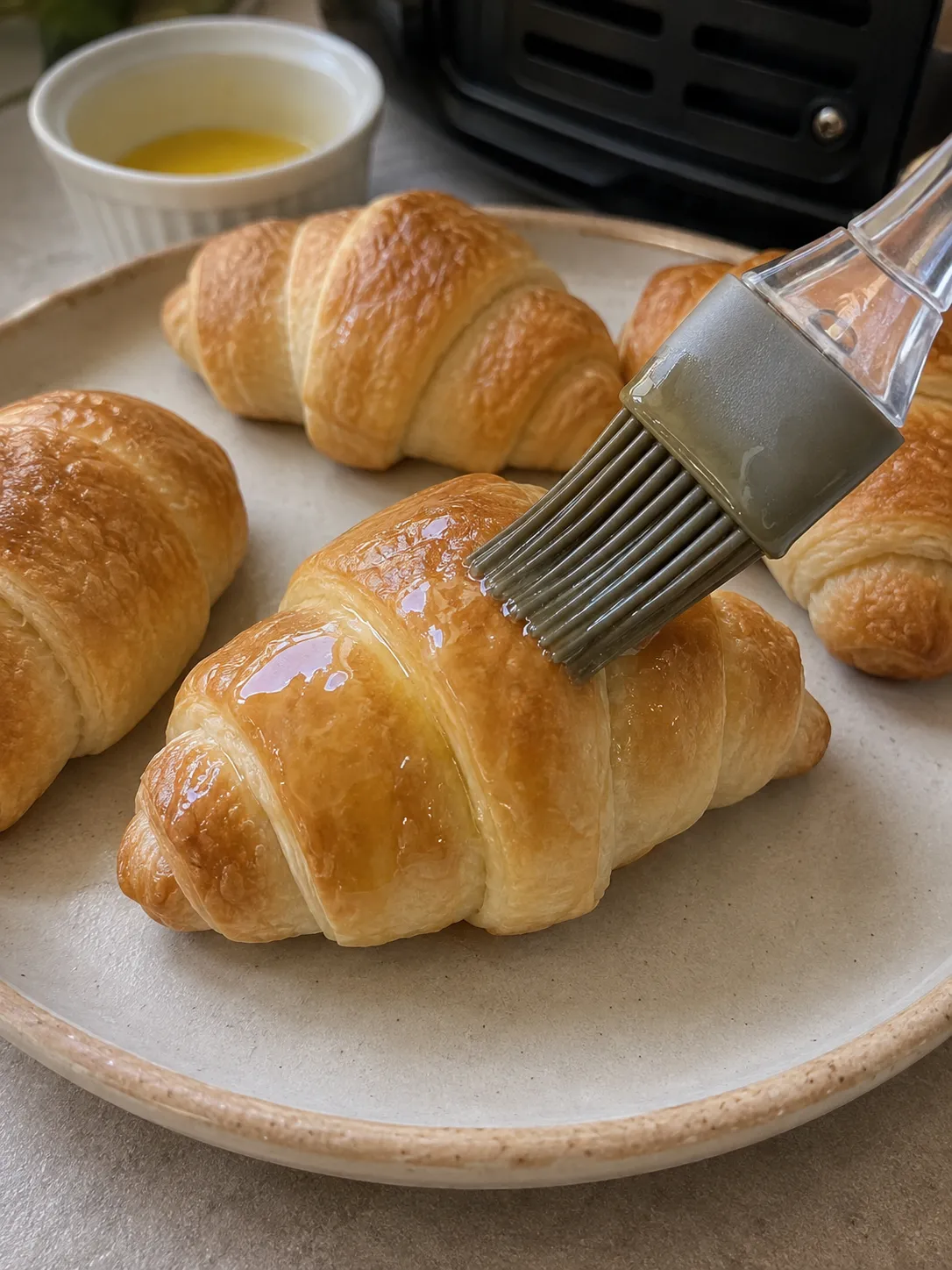 A pastry brush painting melted butter over baked golden crescent rolls on a ceramic plate