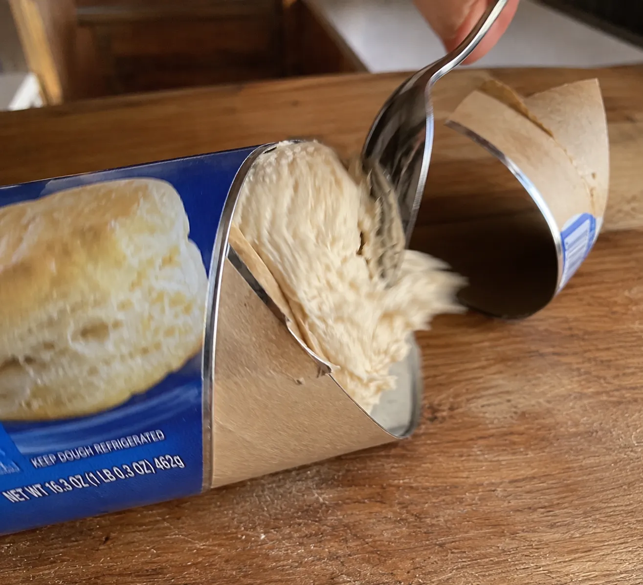 A spoon pressed against the seam of a biscuit can, the dough bursting out through the split