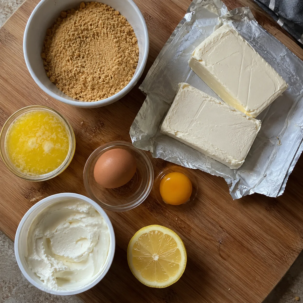 Cheesecake bar ingredients on a wooden board: graham crumbs, melted butter, cream cheese blocks, sour cream, an egg, an egg yolk and half a lemon