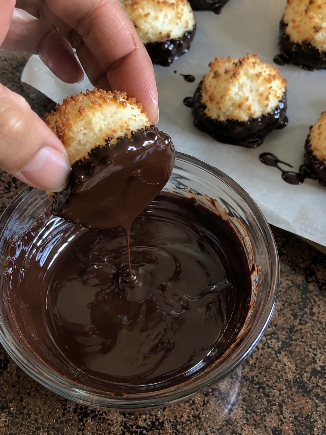 A cooled coconut macaroon being dipped bottom first into a bowl of melted dark chocolate